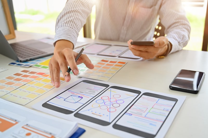 A designer holding a phone While working with UI illustraations on large placed in front of him